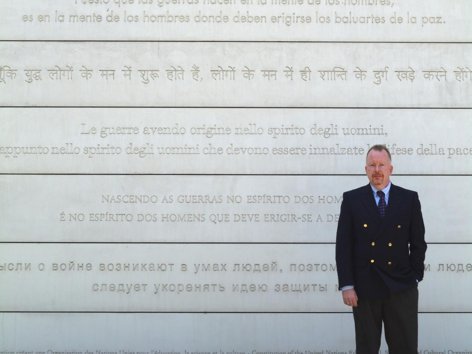 A man in front of the UNESCO Peace Wall with writing in various languages